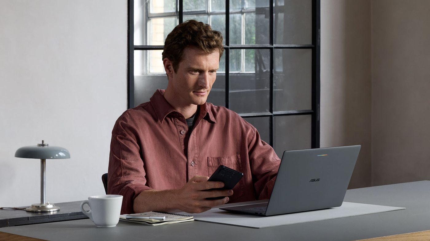 Person at a desk with a closed gray ASUS laptop and a phone in hand