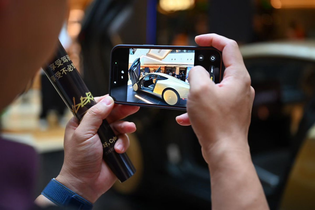 A visitor photographing a Cybercab display on a phone while holding a Tesla souvenir