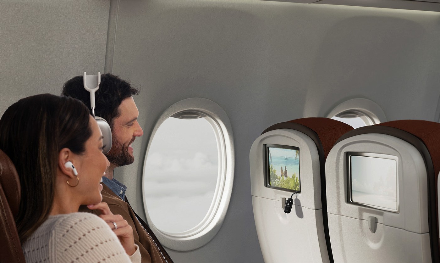Two airplane passengers share one Airfly View plugged into a seatback screen, one in earbuds and one in over-ear headphones