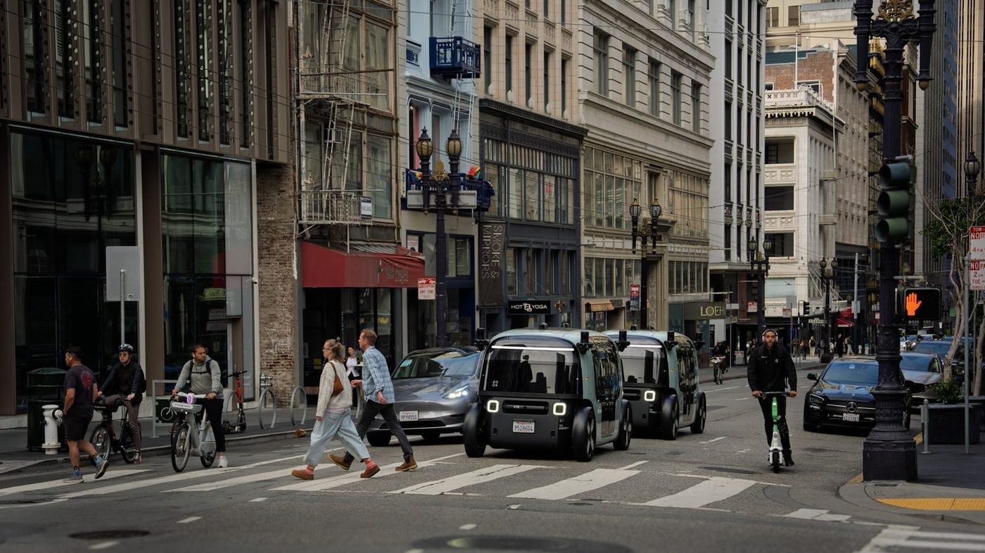 Two Zoox robotaxis in city traffic with pedestrians and cyclists